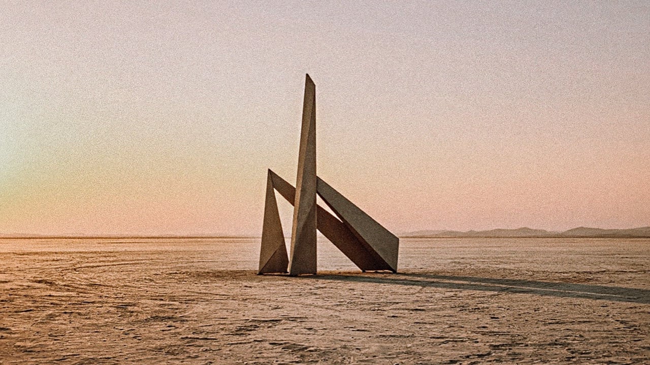 Angular concrete sculpture standing alone on a flat desert lakebed at dusk, casting a long shadow