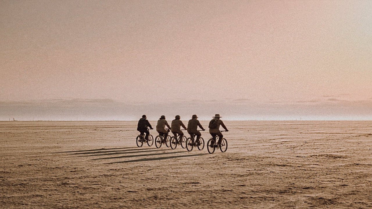 Five people riding bicycles in a line across a flat desert lakebed under a hazy pink sky