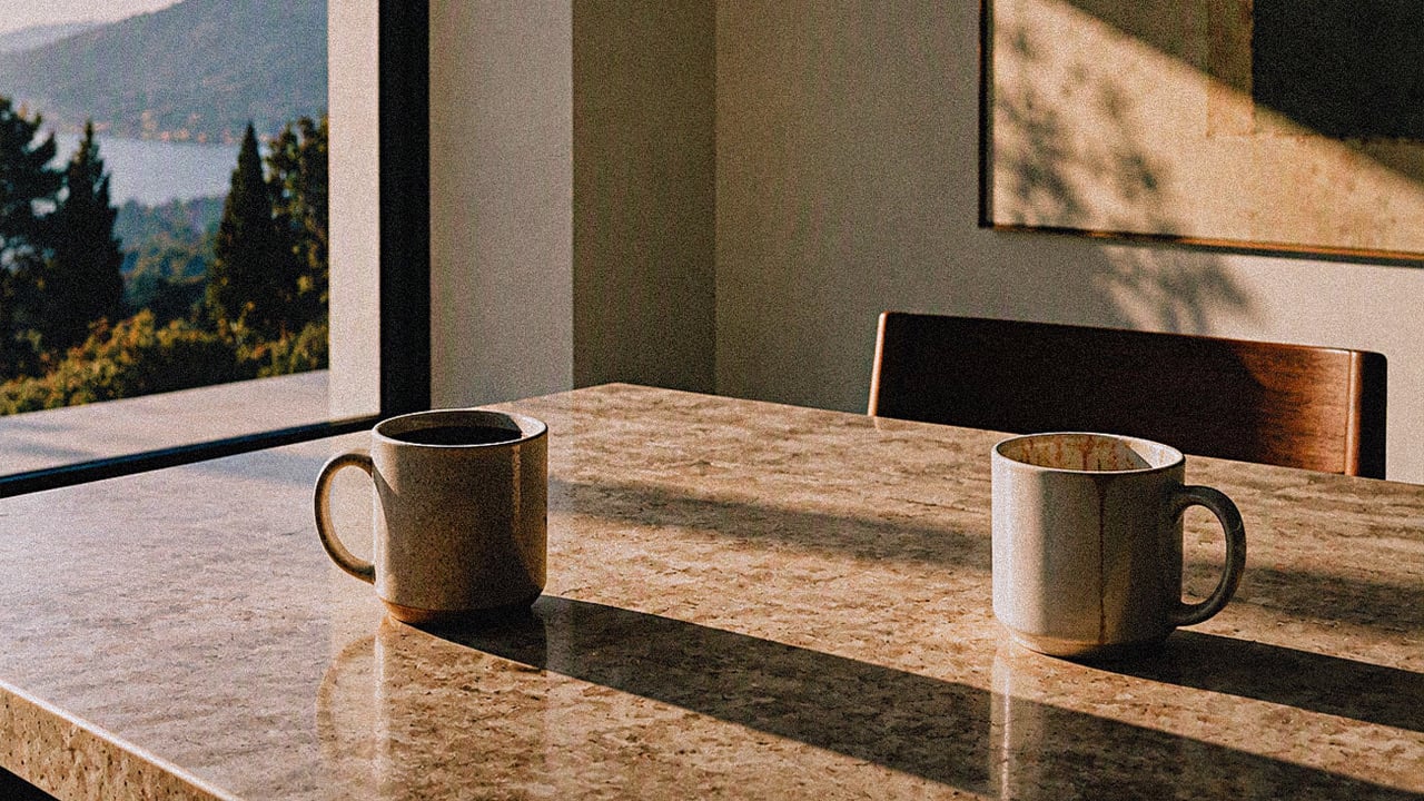 Two coffee mugs on a marble table in morning light, by a window looking out over a lake and wooded hills