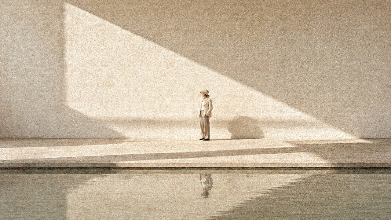 Woman in a sun hat and long linen dress standing alone at the edge of a still pool, facing a pale wall crossed by sunlight and shadow